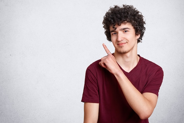 Portret elegancki przystojny młody nastolatek odizolowywający na bielu. Ciemnowłosy kędzierzawy uczeń uśmiecha się i wskazuje na bok