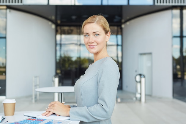 Portret Eleganckiej Blond Bizneswoman Uśmiecha Się Do Kamery Stojąc Przy Stoliku Kawiarnianym Na Lotnisku W Pasie