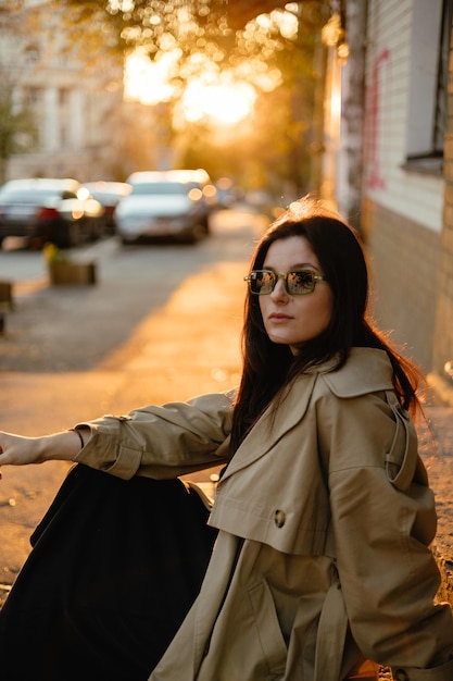 portret eleganckiej dziewczyny w beżowym płaszczu i okularach przeciwsłonecznych na tle ulic jesiennego miasta oświetlonych wieczornym słońcem