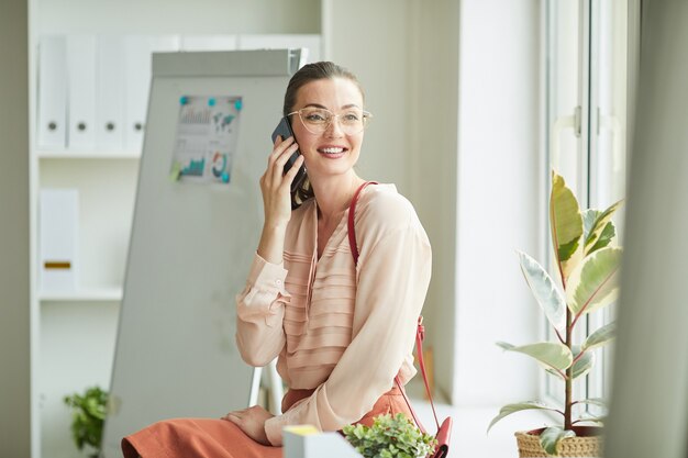 Portret Eleganckiej Młodej Kobiety Mówiącej Przez Telefon, Siedząc Przy Oknie W Biurze I Uśmiechając Się Radośnie