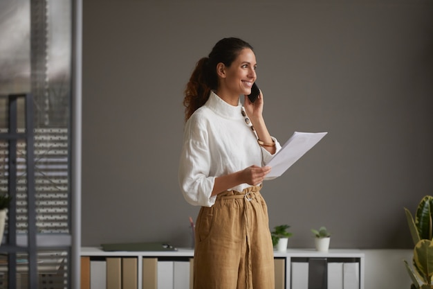 Portret eleganckiej sukcesy bizneswoman mówiąc przez smartfona i uśmiechając się radośnie stojąc przed szarej ścianie w biurze, kopia przestrzeń