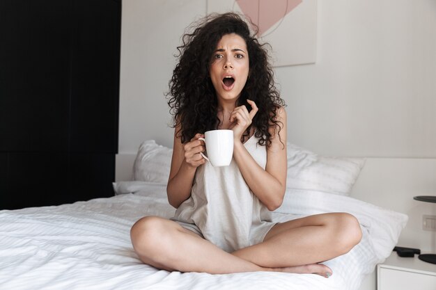 Portret emocjonalnej kręcone kobiety z otwartymi ustami, siedząc na łóżku z białą czystą pościelą w domu rano i trzymając kubek herbaty