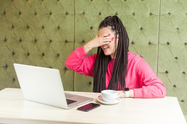 Portret emocjonalnej przestraszonej lub zszokowanej młodej bizneswoman z fryzurą z czarnymi dredami w różowej bluzce siedzącej w kawiarni i zasłaniającej oczy i nie chce patrzeć na ekran laptopa Kryty