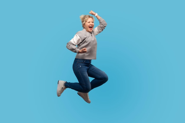 Portret entuzjastycznej szczęśliwej kobiety z krótkimi kręconymi włosami w bluzie skaczącej w powietrzu pokazującej tak, zrobiłem to gest życia energii i koncepcji wolności kryty studio strzał na białym tle na niebieskim tle