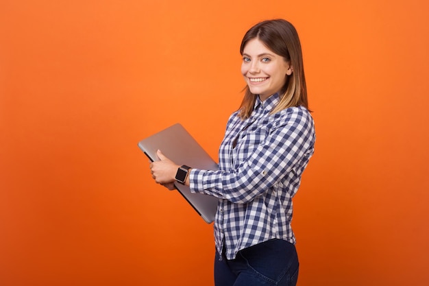 Portret fantazyjnej brunetki z uroczym uśmiechem w kraciastej koszuli na co dzień, stojącej trzymając zamkniętego laptopa i patrząc na kamerę z ząbkowanym uśmiechem w pomieszczeniu studio strzał na białym tle na pomarańczowym tle