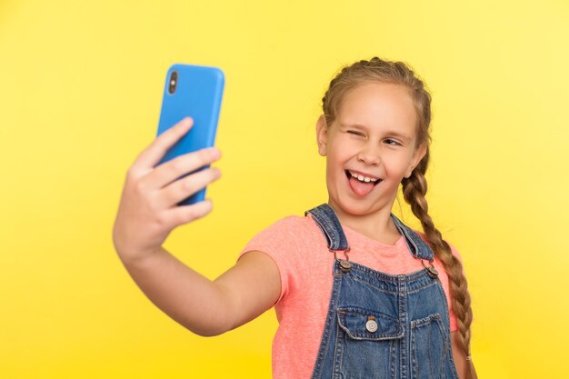 Portret figlarnej śmiesznej dziewczynki z warkoczem w dżinsowych kombinezonach, mrugając i pokazując język podczas robienia selfie dziecka, co twarze rozmawiają na wideorozmowie w pomieszczeniu studio strzał żółte tło