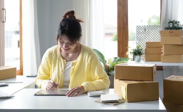 Portret firmy Azjatycka kobieta uśmiecha się i używa tabletu, sprawdzając informacje w pudełku wysyłkowym paczki przed wysłaniem do klienta Przedsiębiorca mała firma pracująca w domu MŚP Biznes online marketing