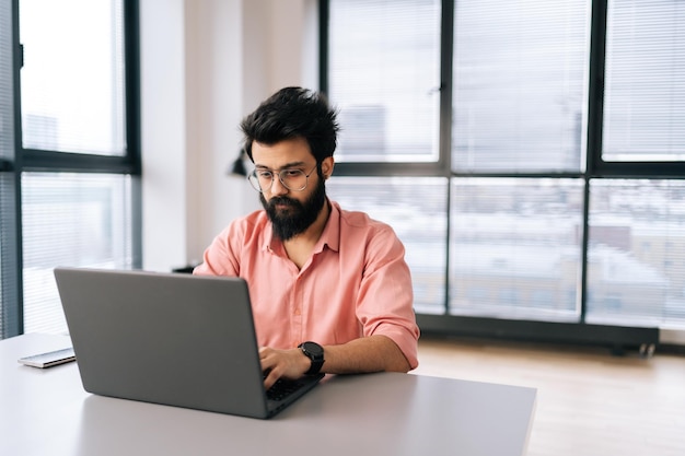 Portret indyjskiego freelancera w przypadkowych ubraniach, pracującego na laptopie, siedzącego przy stole w jasnym biurze coworkingowym na tle okna Brodaty biznesmen w okularach patrzący na ekran komputera