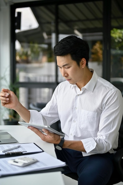 Portret inteligentnego azjatyckiego biznesmena używającego tabletu do zarządzania pracą przy biurku
