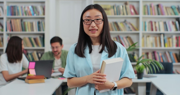 Portret inteligentnej pięknej młodej dziewczyny azjatyckiej w okularach trzymającej książkę w tle bibliotecznej półki na książki, patrząc na kamerę Koncepcja edukacji uniwersyteckiej