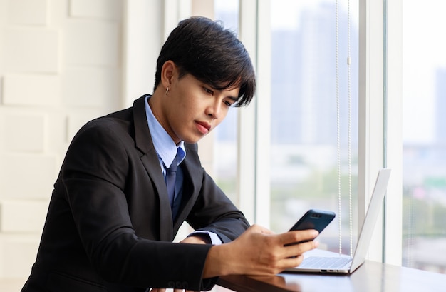 Portret jednego młodego azjatyckiego biznesmena jest inteligentny i przystojny, ubrany w czarny garnitur, siedzący na krześle, rozmawiający przez telefon, a jednocześnie czuć się pewnie z uśmiechem na twarzy patrząc na kamerę z laptopem.