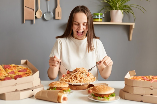 Portret, jeśli jest zdumiony, podekscytowany, bardzo głodna kobieta ubrana w białą koszulkę, siedząca przy stole w kuchni, dostarczająca mnóstwo dań z jedzeniem, jedząca makaron, wyrażająca szczęście