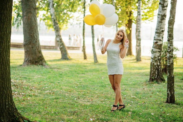 Portret jest ubranym na biel sukni z balonami przy rękami blondynki kobieta przeciw parkowi przy kurnym przyjęciem.