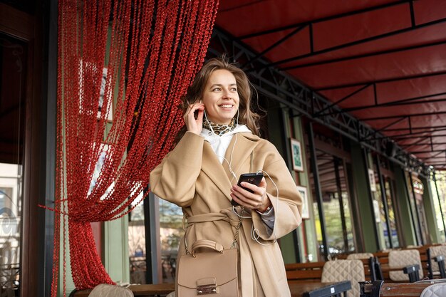 Portret jest ubranym żakiet słucha muzyka na cukiernianej werandzie z słuchawkami trzyma telefon komórkowego w rękach młoda dziewczyna