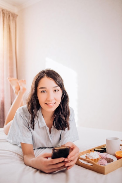 Portret kaukaski atrakcyjna dziewczyna z telefonem, leżąc na łóżku rano w nowoczesnej sypialni. Kobieta uśmiechając się, ciesząc się śniadaniem w łóżku w swoim nowym mieszkaniu. Dzień dobry pozytywne emocje. Skopiuj miejsce
