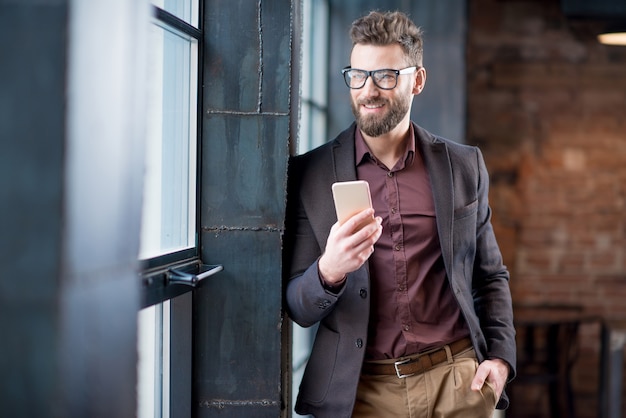 Portret kaukaski brodaty biznesmen ubrany w garnitur z okularami, trzymając inteligentny telefon w pobliżu okna w biurze wewnętrznym na poddaszu