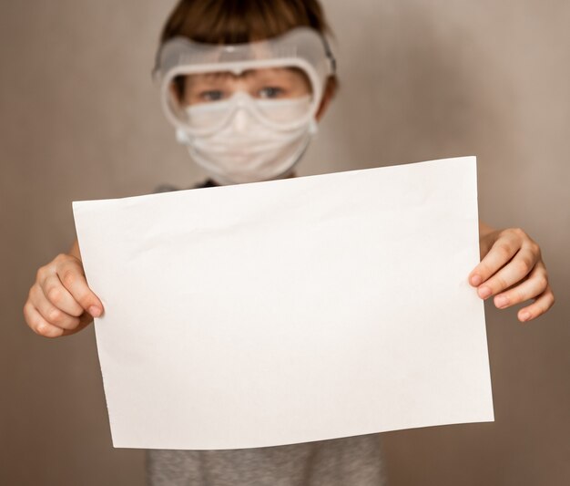 Portret kaukaski chłopiec w masce ochronnej respiratora trzyma pustą kartkę papieru na szarym tle. Ochrona przed koronawirusem. Makieta, miejsce na kopię, reklama