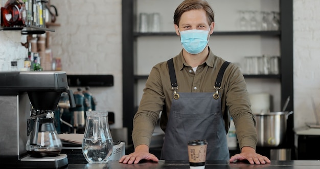 Portret kaukaski przystojny mężczyzna barista w masce medycznej i rękawiczkach, dając filiżankę kawy do aparatu i uśmiechając się. Barista wręczający napój kawa na wynos w kawiarni.