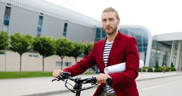 Portret kaukaski przystojny młody człowiek w czerwonej kurtce stojący na zewnątrz z rowerem lub skuter elektryczny i patrząc na kamery. Przystojny stylowy facet na rowerze trzymając papier lub rysując na ulicy miasta