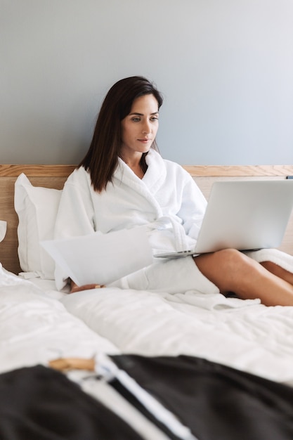 Portret kaukaskiej dorosłej bizneswoman w białym szlafroku pracującym z papierowymi dokumentami i laptopem, leżąc na łóżku w mieszkaniu