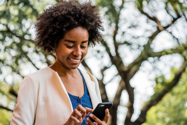 Portret kobiety biznesu afro przy użyciu swojego telefonu komórkowego, stojąc na zewnątrz w parku