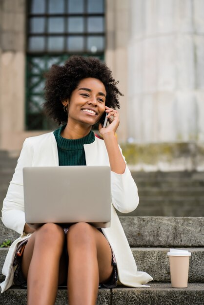 Portret Kobiety Biznesu Afro Rozmawia Przez Telefon I Przy Użyciu Swojego Laptopa, Siedząc Na Schodach Na Zewnątrz