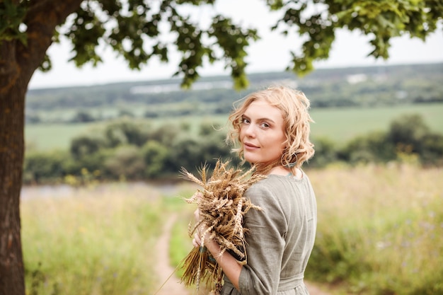 Portret Kobiety Blondynka Ubrana W Naturalną Lnianą Sukienkę Z Bukietem Pszenicy W Dłoniach W Lecie Na świeżym Powietrzu.