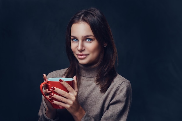 Portret kobiety brunetka ubrana w brązową kurtkę z długą szyją trzyma filiżankę czerwonej kawy.