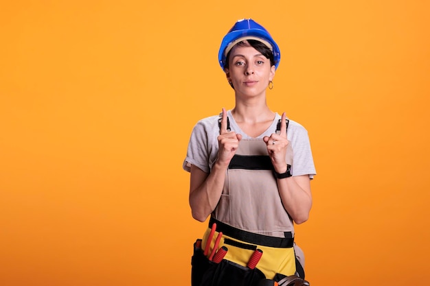 Zdjęcie portret kobiety budowniczej wskazującej w górę palcami wskazującymi w górę i wskazujący kierunek w górę. pozuje w studiu na żółtym tle i nosi kapelusz z kombinezonem.