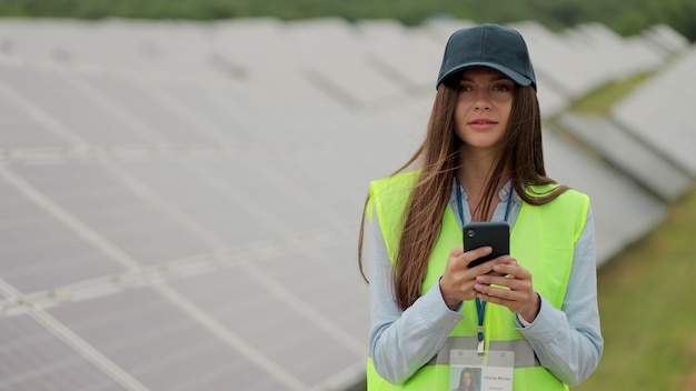 Portret kobiety inżynier inspektor w mundurze używa smartfona na elektrowni słonecznej. Pracownik farmy słonecznej stojący w pobliżu rzędów z bateriami. Pole paneli słonecznych. Produkcja czystej energii. Zielona energia.