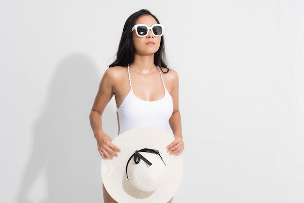Portret kobiety lato studio. Azjatycka kobieta ubrana w biały strój kąpielowy w pozycji stojącej, trzymająca biały biały kapelusz i okulary przeciwsłoneczne, w letni sposób na odosobnionym białym tle.