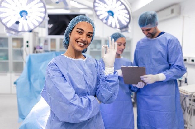 Portret kobiety pielęgniarki chirurga LUB członka personelu ubranego w fartuch chirurgiczny, maskę i siatkę do włosów w szpitalnej sali operacyjnej, nawiązując kontakt wzrokowy, uśmiechając się, zadowolony, szczęśliwy, patrząc na kamerę