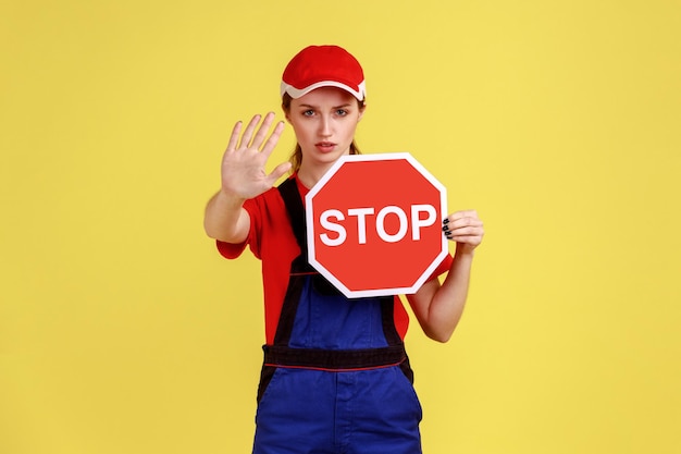 Portret kobiety robotnicy pokazujący gest zatrzymania i trzymający czerwony znak drogowy, zakazy i ograniczenia, ubrany w kombinezon i czerwoną czapkę. Kryty studio strzał na białym tle na żółtym tle.