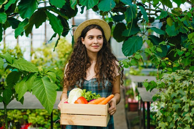 Portret kobiety rolnika spacerującej w szklarni z pudełkiem organicznych warzyw