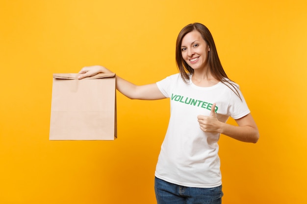 Portret kobiety w biały t-shirt napisany napis zielony tytuł wolontariusz trzymać puste rzemiosło papierową torbę na wynos na białym tle na żółtym tle. Dobrowolna bezpłatna pomoc pomaga w koncepcji łaski charytatywnej