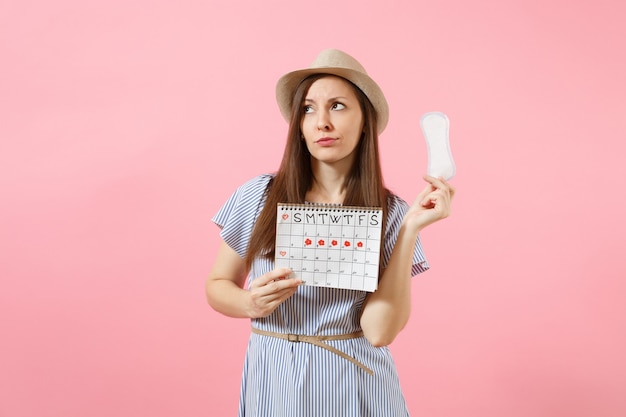 Portret kobiety w niebieskiej sukience, kapeluszu trzymając podpaskę, kalendarz kobiet okresów do sprawdzania dni menstruacji na białym tle na różowym tle. Medycyna, opieka zdrowotna, koncepcja ginekologiczna. Skopiuj miejsce