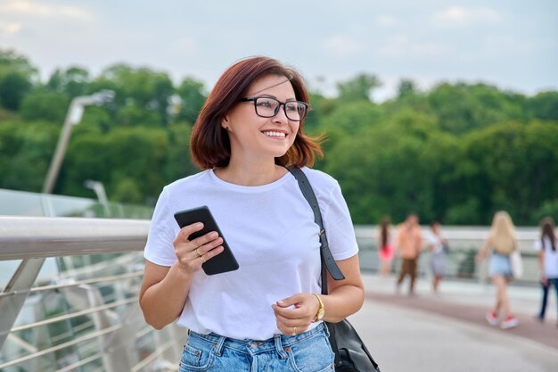 Portret kobiety w średnim wieku spacerującej ze smartfonem w ręku letni dzień w mieście