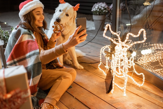 Portret kobiety w świątecznym kapeluszu i kracie ze swoim uroczym psem świętującym święta Nowego Roku w domu, karmiącym psa piernikowymi ciasteczkami i robiącym zdjęcie selfie