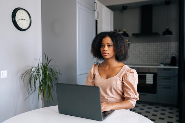 Portret kręconej afrykańskiej freelancerki pracującej lub studiującej zdalnie z domowego biura przy użyciu laptopa przy biurku w nowoczesnej kuchni patrzącej na kamerę