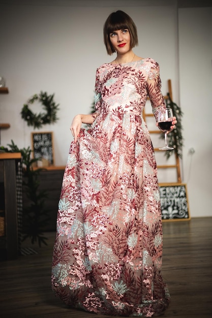 Portret kryty pewna siebie dziewczyna w długiej sukni pozowanie w pobliżu choinki z lampką wina. Nieostrość. Selektywne skupienie.