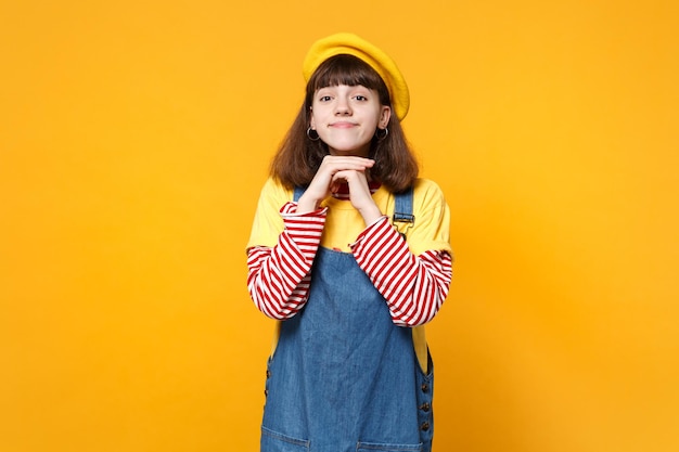 Portret ładna dziewczyna nastolatka w francuski beret, dżinsowa sukienka trzymając się za ręce w pobliżu twarzy na białym tle na tle żółtej ściany w studio. Ludzie szczere emocje, koncepcja stylu życia. Makieta miejsca na kopię.