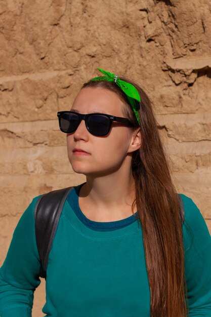 Portret ładnej dziewczyny hipster w okularach przeciwsłonecznych, zielonej bluzie z chustką, stojącej na tle glinianej skały w słoneczny dzień