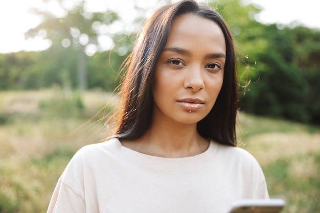 Portret ładnej kobiety noszącej piercing do ust, patrzącej na kamerę i trzymającej smartfon w zielonym parku