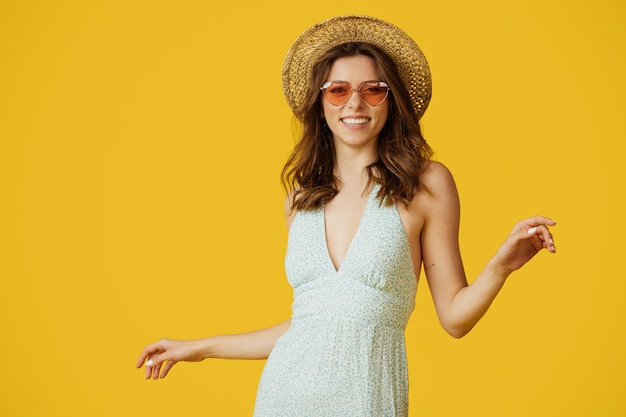 Portret ładnej kobiety w letniej sukience słomkowy kapelusz i kolorowe okulary przeciwsłoneczne pozowanie i taniec na żółtym tle