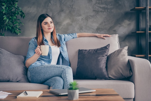 Portret ładnej, ładnej, zadowolonej dziewczyny, siedzącej na kanapie, trzymającej kubek cappuccino