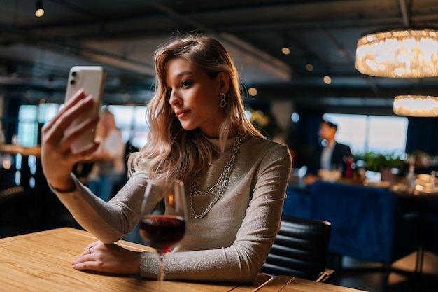 Portret ładnej młodej kobiety siedzącej przy stole z kieliszkiem czerwonego wina za pomocą telefonu komórkowego na żywo w mediach społecznościowych dla biznesu, pisząc historie robiąc selfie