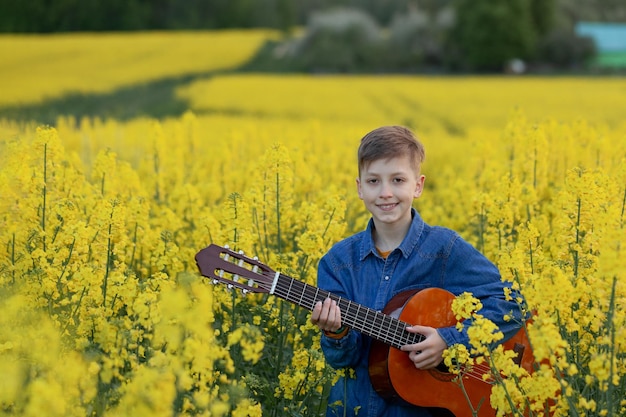 Portret ładny chłopiec gra na gitarze w letnim polu żółtym. Portret uśmiechniętego dziecka