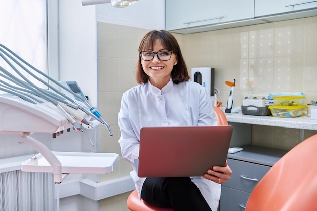 Portret lekarza dentystycznego siedzącego w biurze z laptopem w rękach patrzącym na kamerę Dojrzała uśmiechnięta pielęgniarka w klinice stomatologicznej Medycyna stomatologiczna