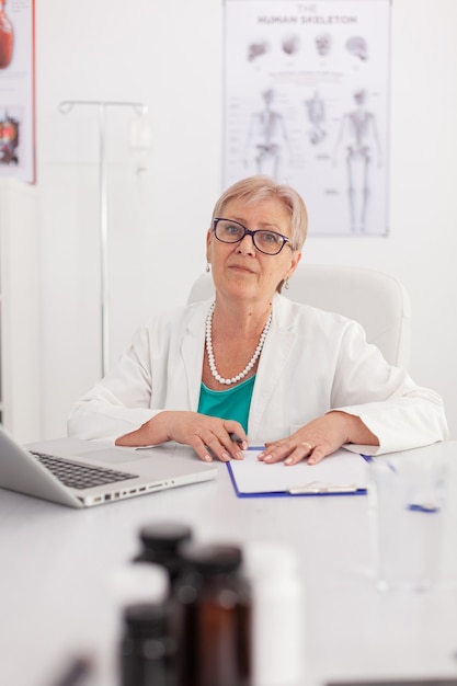 Zdjęcie portret lekarza senior kobieta lekarz patrząc w kamerę siedzi przy biurku w sali konferencyjnej pracy w leczeniu tabletek chorobowych. pewny siebie lekarz przedstawiający wiedzę medyczną