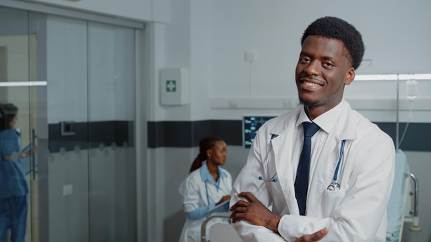 Portret lekarza z stetoskopem stojący na oddziale szpitalnym. Specjalista o białym fartuchu i skrzyżowanych ramionach leczący kobietę z chorobą. Lekarz ogólny uśmiecha się i patrzy w kamerę